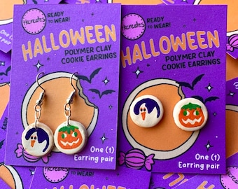 May include: Two pairs of Halloween polymer clay cookie earrings on purple display cards. One pair features dangling charms with a ghost and a jack-o'-lantern. The other pair features stud earrings with a ghost and a jack-o'-lantern design.