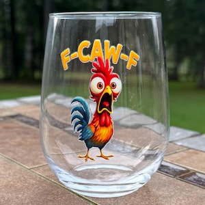 May include: A clear stemless wine glass with a cartoon rooster design. The rooster is red, orange, yellow, and blue with a wide open mouth and large eyes. The text "F-CAW-F" is above the rooster in yellow letters.