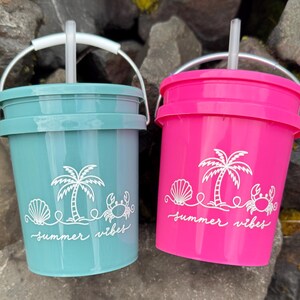 May include: Two plastic bucket-shaped tumblers with lids and straws. One is teal, the other is hot pink. Both have white graphics of a palm tree, seashell, and crab, with the words "summer vibes". The tumblers have silver handles.