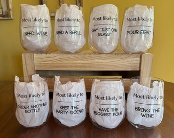 May include: Eight clear stemless wine glasses, each filled with white tissue paper. Each glass features black text reading Most likely to followed by unique phrases including NEED WINE, NEED A REFILL, SAY JUST ONE GLASS, POUR FIRST, ORDER ANOTHER BOTTLE, KEEP THE PARTY GOING, HAVE THE BIGGEST POUR, and BRING THE WINE.