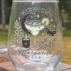 May include: A clear stemless glass featuring a printed graphic of a margarita, a crescent moon, a butterfly, a candle, and olive branches. The text reads MIDNIGHT MARGARITA CLUB. The design includes gold star accents and a lime slice garnish.