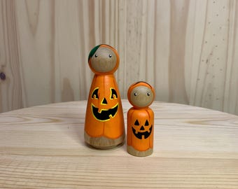 May include: Two wooden peg dolls dressed as pumpkins. The larger doll is orange with a black jack-o'-lantern face and a green and brown hat. The smaller doll has a similar design. Both dolls have a natural wood base.