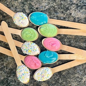 Puede incluir: Cucharas de madera con coberturas coloridas. Algunas cucharas tienen glaseado blanco con chispas coloridas, mientras que otras tienen glaseado azul, verde y rosa. Las cucharas están dispuestas sobre una superficie oscura y texturizada.