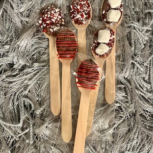 Puede incluir: Cucharas de madera cubiertas con chocolate y varios ingredientes, incluyendo menta triturada, goteo rojo y malvaviscos. Las cucharas est&aacute;n envueltas individualmente y dispuestas sobre una superficie gris texturizada.