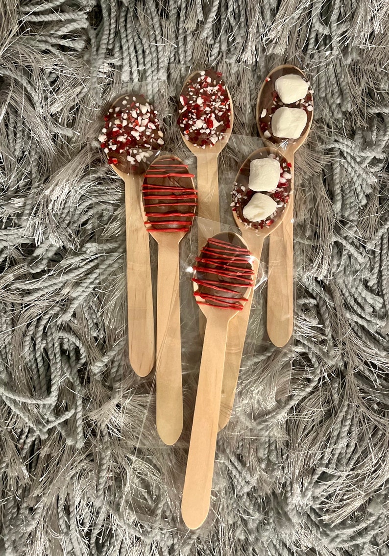 Puede incluir: Cucharas de madera cubiertas con chocolate y varios ingredientes, incluyendo menta triturada, goteo rojo y malvaviscos. Las cucharas est&aacute;n envueltas individualmente y dispuestas sobre una superficie gris texturizada.