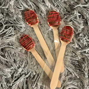 Puede incluir: Cuatro cucharas de madera, cada una cubierta con un &oacute;valo de chocolate y un glaseado rojo, est&aacute;n empaquetadas en pl&aacute;stico transparente. Las cucharas est&aacute;n dispuestas sobre una superficie gris texturizada.