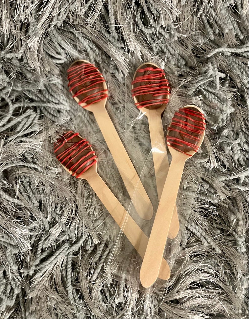 Puede incluir: Cuatro cucharas de madera, cada una cubierta con un &oacute;valo de chocolate y un glaseado rojo, est&aacute;n empaquetadas en pl&aacute;stico transparente. Las cucharas est&aacute;n dispuestas sobre una superficie gris texturizada.