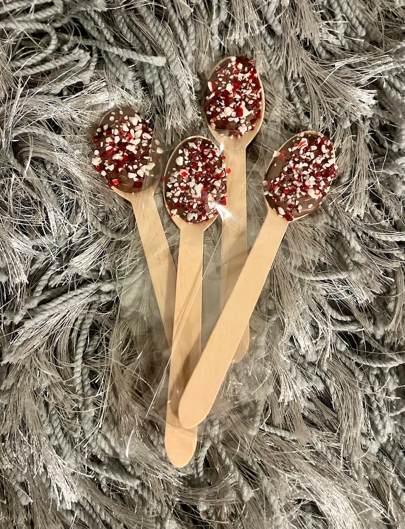 Puede incluir: Cuatro cucharas de madera con chocolate y chispas rojas y blancas. Las cucharas est&aacute;n dispuestas en una bolsa de pl&aacute;stico transparente sobre una superficie gris texturizada. El chocolate cubre la cuchara.