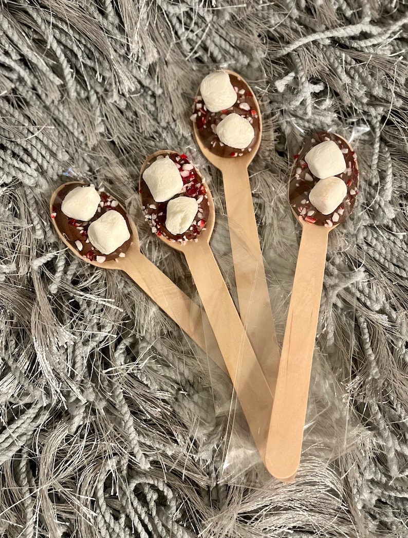 Puede incluir: Cuatro cucharas de madera con chocolate, malvaviscos y caramelos de menta triturados. Est&aacute;n empaquetadas en una bolsa de pl&aacute;stico transparente, sobre una superficie gris texturizada. Para chocolate caliente.