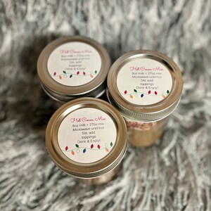 Puede incluir: Tres frascos de vidrio con tapas plateadas, cada uno etiquetado "Hot Cocoa Mix". Las etiquetas incluyen instrucciones y una cadena de luces de colores. Los frascos est&aacute;n dispuestos sobre una superficie suave, gris y texturizada.