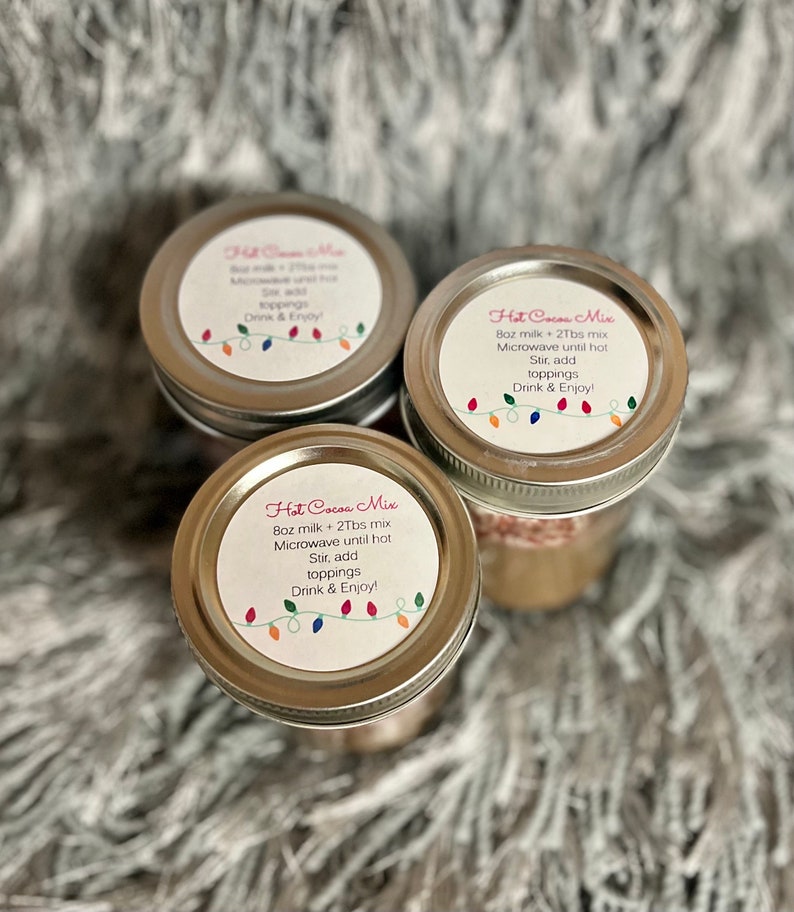 Puede incluir: Tres frascos de vidrio con tapas plateadas, cada uno etiquetado "Hot Cocoa Mix". Las etiquetas incluyen instrucciones y una cadena de luces de colores. Los frascos est&aacute;n dispuestos sobre una superficie suave, gris y texturizada.