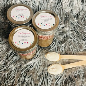 Puede incluir: Tres tarros de mezcla para chocolate caliente con tapas, cada uno etiquetado con instrucciones y una cadena de luces de colores. Dos cucharas de madera con sustancia blanca est&aacute;n en primer plano. Los tarros est&aacute;n sobre una superficie gris y esponjosa.
