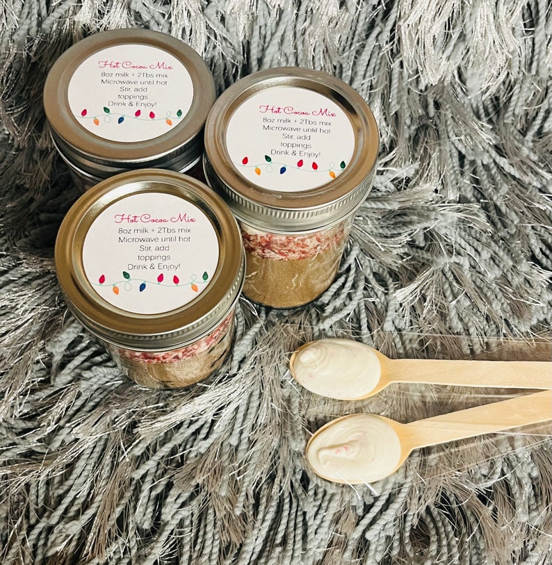 Puede incluir: Tres tarros de mezcla para chocolate caliente con tapas, cada uno etiquetado con instrucciones y una cadena de luces de colores. Dos cucharas de madera con sustancia blanca est&aacute;n en primer plano. Los tarros est&aacute;n sobre una superficie gris y esponjosa.