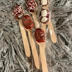 Puede incluir: Un conjunto de seis cucharas de madera, cada una cubierta de chocolate y adornada con diferentes ingredientes. Algunas tienen chispas rojas y blancas, otras malvaviscos y algunas tienen un glaseado rojo. Las cucharas est&aacute;n empaquetadas en pl&aacute;stico transparente.