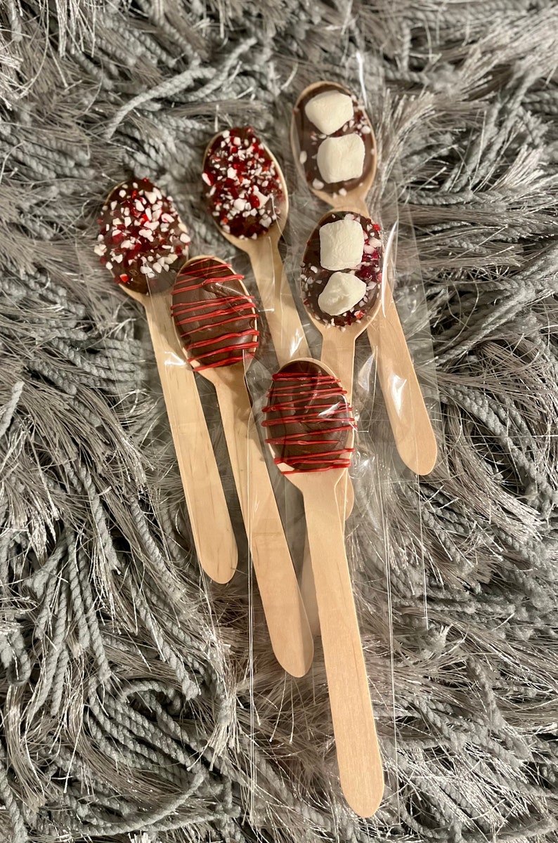 Puede incluir: Un conjunto de seis cucharas de madera, cada una cubierta de chocolate y adornada con diferentes ingredientes. Algunas tienen chispas rojas y blancas, otras malvaviscos y algunas tienen un glaseado rojo. Las cucharas est&aacute;n empaquetadas en pl&aacute;stico transparente.