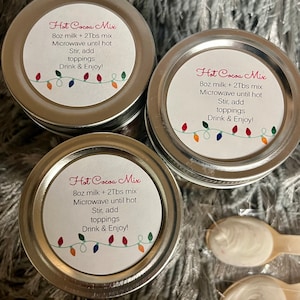 Puede incluir: Tres peque&ntilde;os frascos con tapa plateada de mezcla para chocolate caliente con etiquetas que dicen "Hot Cocoa Mix". Las etiquetas tambi&eacute;n incluyen instrucciones. Dos peque&ntilde;as cucharas de madera con sustancia blanca est&aacute;n en la imagen. Los frascos est&aacute;n sobre una superficie gris y difusa.