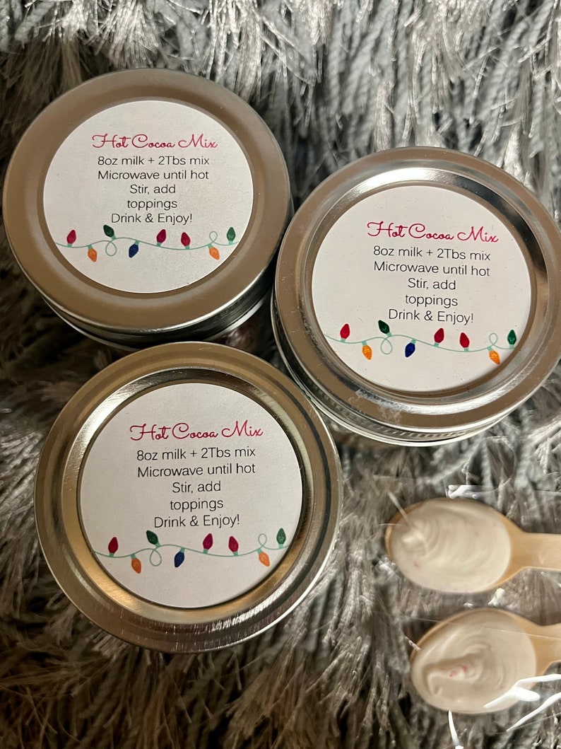 Puede incluir: Tres peque&ntilde;os frascos con tapa plateada de mezcla para chocolate caliente con etiquetas que dicen "Hot Cocoa Mix". Las etiquetas tambi&eacute;n incluyen instrucciones. Dos peque&ntilde;as cucharas de madera con sustancia blanca est&aacute;n en la imagen. Los frascos est&aacute;n sobre una superficie gris y difusa.