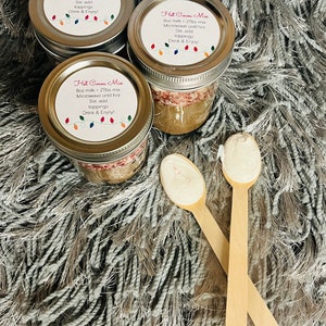 Puede incluir: Tres tarros de cristal de mezcla de chocolate caliente con tapas plateadas, cada uno etiquetado con instrucciones y una cadena de luces de colores. Dos cucharas de madera con cobertura blanca frente a los tarros.