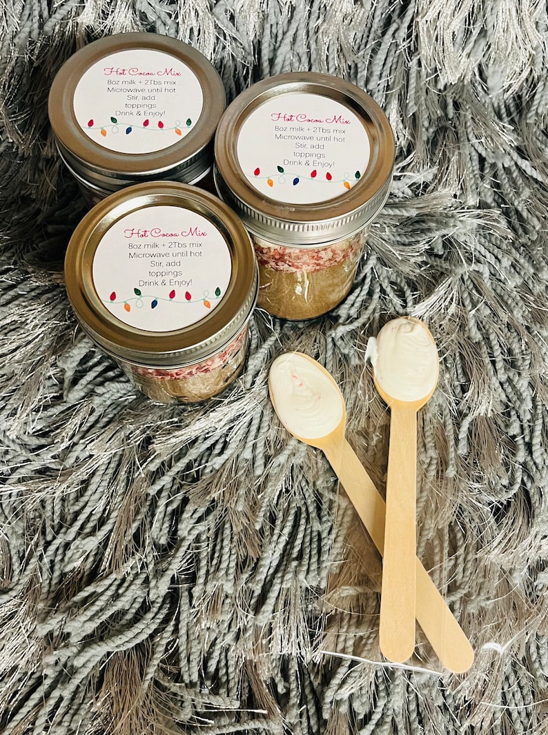Puede incluir: Tres tarros de cristal de mezcla de chocolate caliente con tapas plateadas, cada uno etiquetado con instrucciones y una cadena de luces de colores. Dos cucharas de madera con cobertura blanca frente a los tarros.