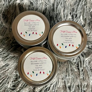 Puede incluir: Tres peque&ntilde;os frascos con tapa plateada de mezcla para chocolate caliente. Cada frasco tiene una etiqueta blanca con las palabras "Hot Cocoa Mix" e instrucciones. Una cadena de luces navide&ntilde;as de colores decora la parte inferior de la etiqueta.