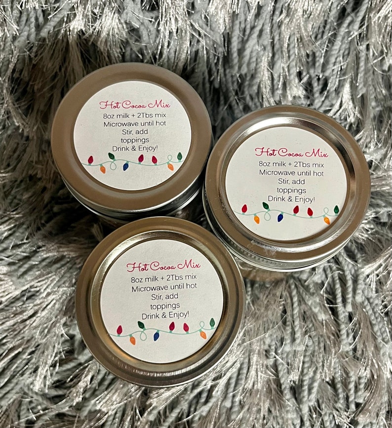 Puede incluir: Tres peque&ntilde;os frascos con tapa plateada de mezcla para chocolate caliente. Cada frasco tiene una etiqueta blanca con las palabras "Hot Cocoa Mix" e instrucciones. Una cadena de luces navide&ntilde;as de colores decora la parte inferior de la etiqueta.