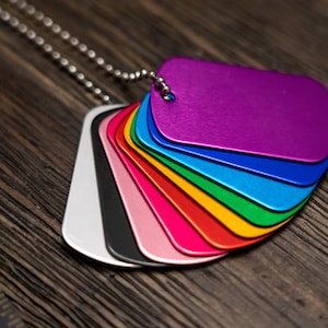 Puede incluir: Un conjunto de chapas de perro de metal de colores en una cadena plateada. Las chapas est&aacute;n dispuestas en forma de arco&iacute;ris, con la chapa superior de color morado y la inferior de color plateado.