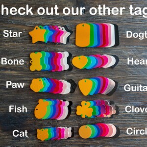 Puede incluir: Surtido de etiquetas met&aacute;licas de colores en varias formas, incluyendo estrella, hueso, pata, pez, gato, coraz&oacute;n, p&uacute;a de guitarra, tr&eacute;bol y c&iacute;rculo. Las etiquetas est&aacute;n dispuestas en filas sobre una superficie de madera. El texto "Check out our other tags!" est&aacute; en la parte superior de la imagen.