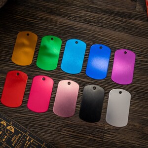 Puede incluir: Diez etiquetas de perro de metal en blanco en varios colores, incluyendo dorado, verde, azul, morado, rojo, rosa, plateado, negro y dos tonos de gris. Las etiquetas est&aacute;n dispuestas en una fila sobre una superficie de madera.