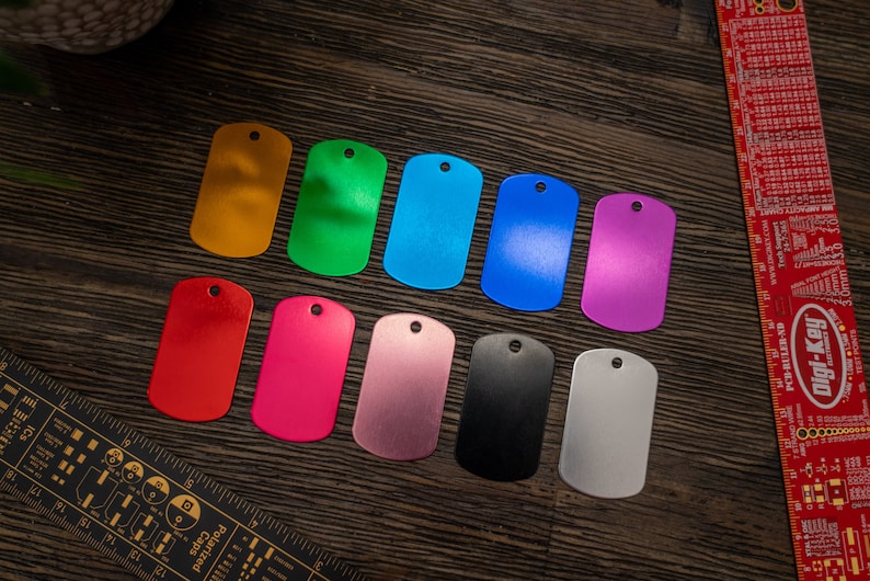 Puede incluir: Diez etiquetas de perro de metal en blanco en varios colores, incluyendo dorado, verde, azul, morado, rojo, rosa, plateado, negro y dos tonos de gris. Las etiquetas est&aacute;n dispuestas en una fila sobre una superficie de madera.