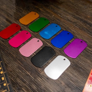 Puede incluir: Un conjunto de diez chapas de perro de metal en blanco en varios colores, incluidos rojo, rosa, naranja, verde, azul, morado, negro y plateado. Las chapas est&aacute;n dispuestas en una fila sobre una superficie de madera.