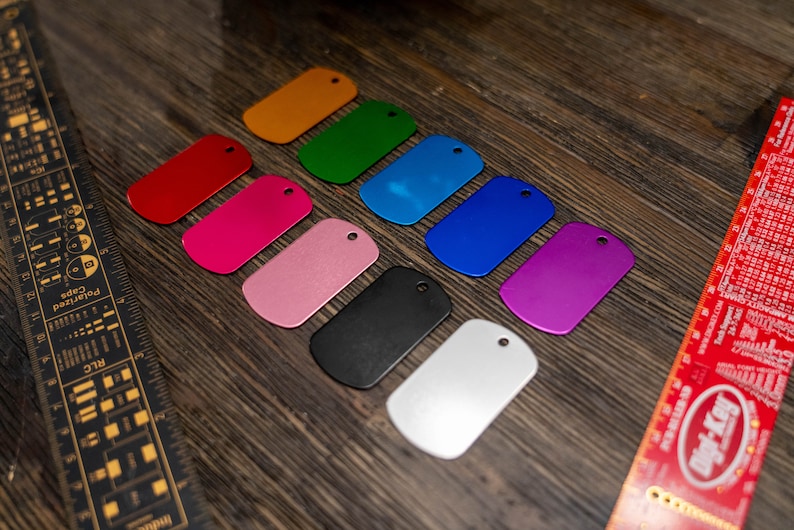 Puede incluir: Un conjunto de diez chapas de perro de metal en blanco en varios colores, incluidos rojo, rosa, naranja, verde, azul, morado, negro y plateado. Las chapas est&aacute;n dispuestas en una fila sobre una superficie de madera.