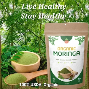 May include: A brown paper bag with a white label that says "Organic Moringa" and "100% Organic Moringa Powder". The bag is sitting on a wooden surface with a green leafy background. The text "Live Healthy Stay Healthy" is at the top of the image. The bag is 100% USDA Organic.