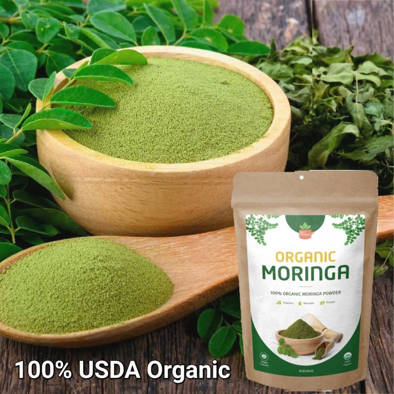May include: A brown paper bag with a label that reads "Organic Moringa 100% Organic Moringa Powder". The bag is filled with green powder. A wooden bowl and spoon are filled with the same green powder. The background is a wooden surface with green leaves.
