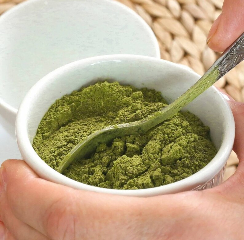 May include: A white ceramic bowl filled with a bright green powder, a silver spoon is partially submerged in the powder.