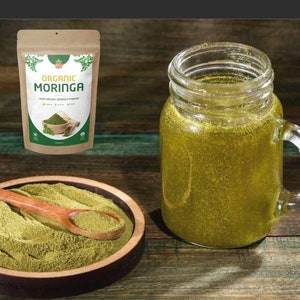 May include: A brown paper bag with a white label that reads "Organic Moringa 100% Organic Moringa Powder" with a green leaf design. A wooden spoon is filled with green powder in a wooden bowl. A glass jar with a handle is filled with a green liquid.