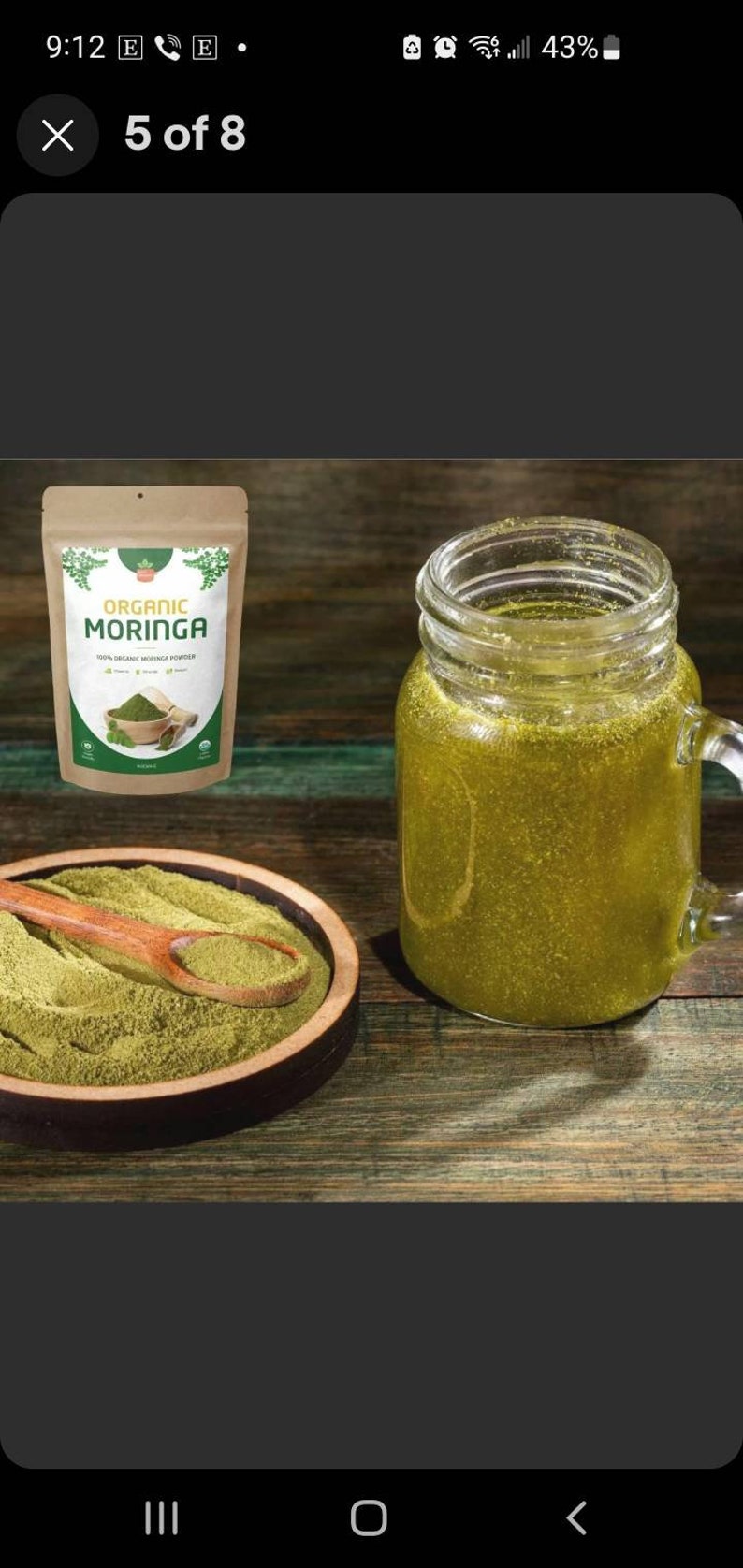 May include: A brown paper bag with a white label that reads "Organic Moringa 100% Organic Moringa Powder" with a green leaf design. A wooden spoon is filled with green powder in a wooden bowl. A glass jar with a handle is filled with a green liquid.