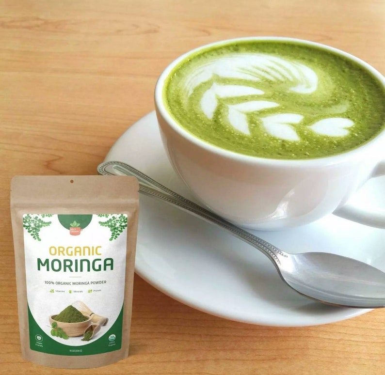 May include: A brown paper bag with a label that reads "Organic Moringa 100% Organic Moringa Powder". The bag is sitting on a wooden table next to a white cup of green tea with latte art.
