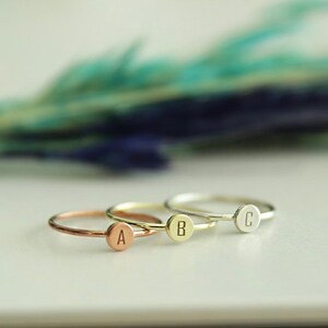 May include: Three delicate rings with a single letter engraved on each. The rings are in different metal finishes: rose gold, gold, and silver. The letters are A, B, and C.