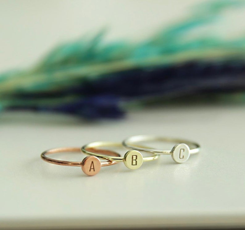 May include: Three delicate rings with a single letter engraved on each. The rings are in different metal finishes: rose gold, gold, and silver. The letters are A, B, and C.