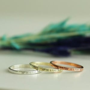 May include: Three thin rings in silver, gold, and rose gold are displayed. Each ring has engraved text with numbers and letters. The rings are on a light surface, with a blurred background of green and blue.