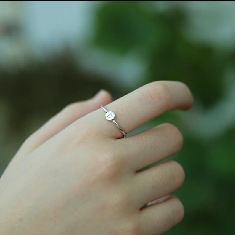 May include: A silver ring with a small round charm featuring the letter "C" on it. The ring is worn on a person's finger.