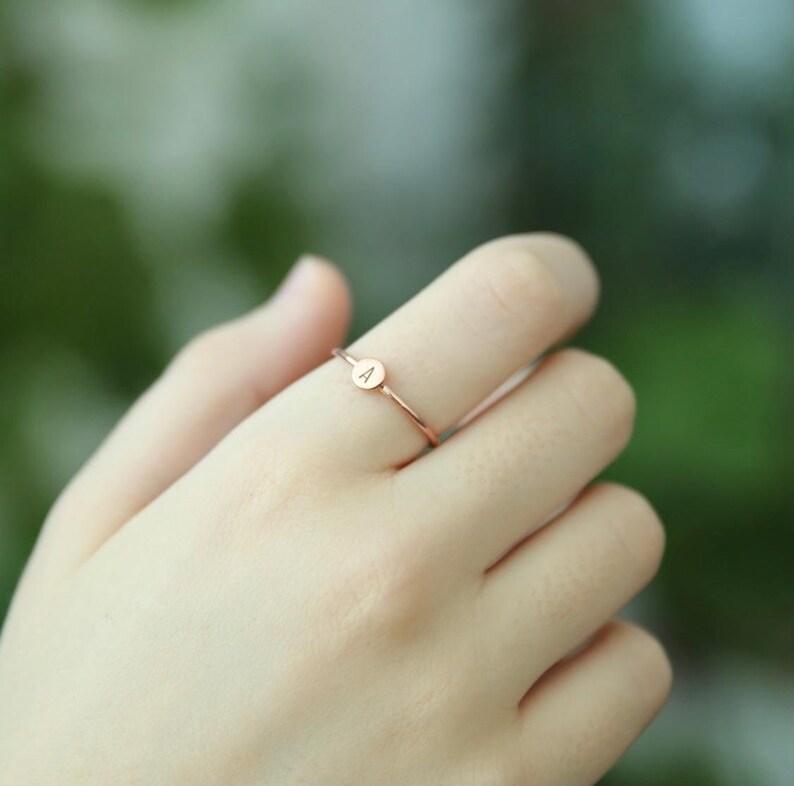 May include: A delicate rose gold ring with a small round disc featuring the letter "A" engraved on it.
