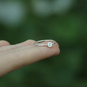 May include: A silver ring with a small, round disc featuring the letter "C" engraved on it.