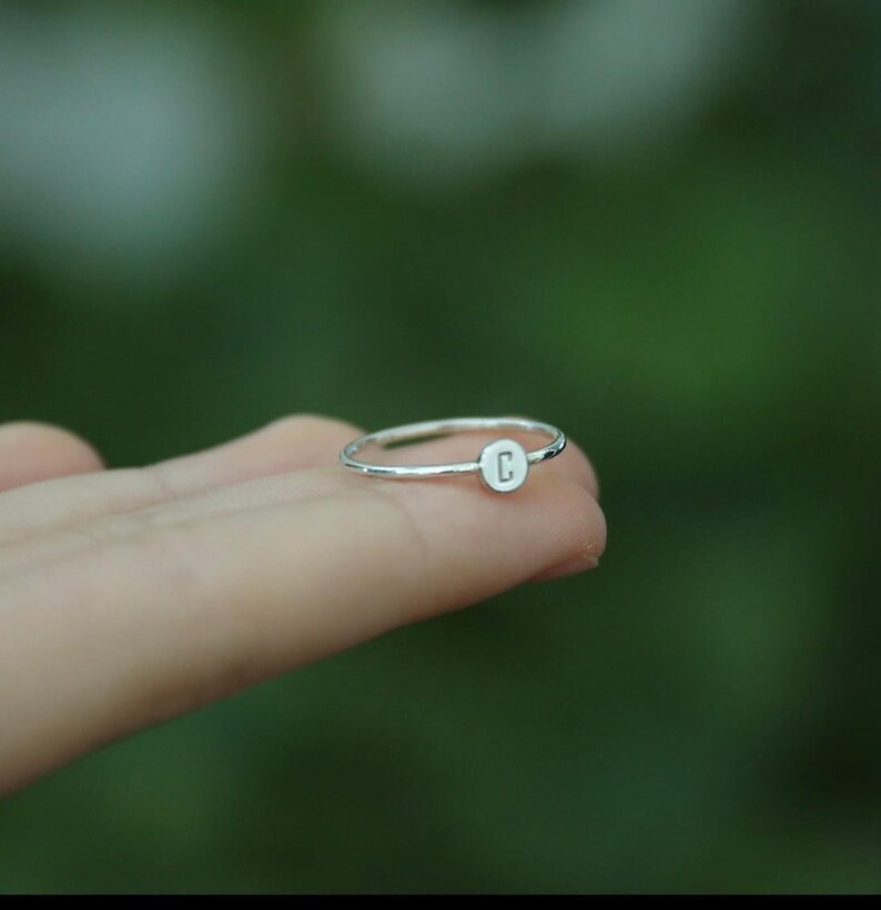 May include: A silver ring with a small, round disc featuring the letter "C" engraved on it.