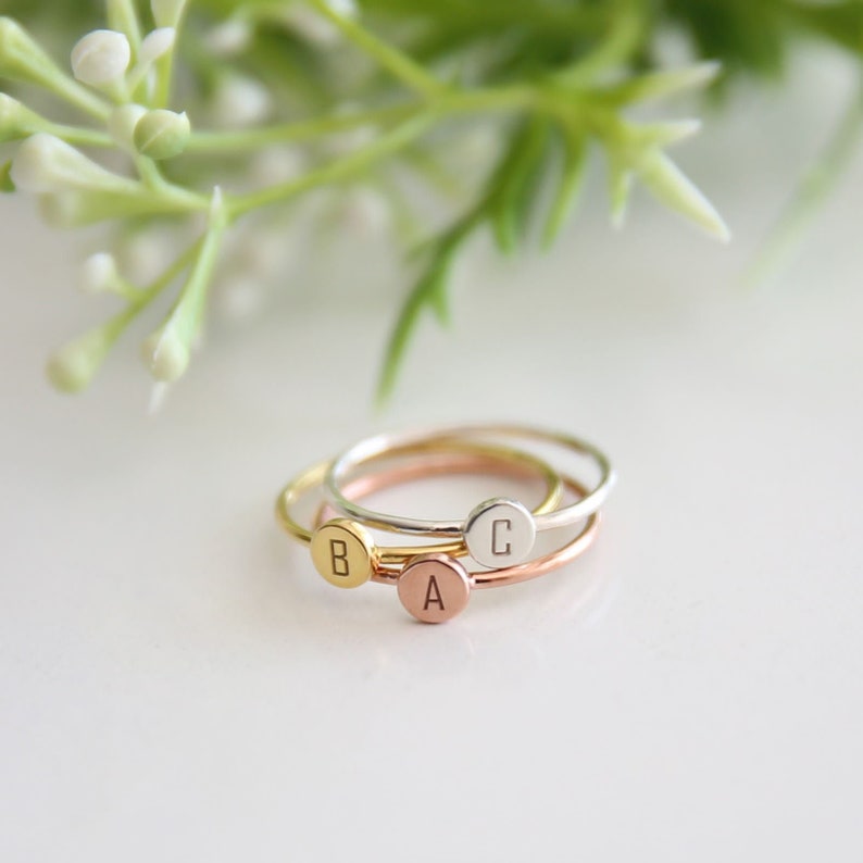 May include: Three delicate stacking rings with a minimalist design. The rings are made of gold, silver, and rose gold and feature a small round disc with a letter engraved on each. The letters are A, B, and C.