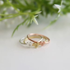 Może przedstawiać: Trzy delikatne pierścionki w kolorze srebrnym, złotym i różowego złota, każdy z zawieszką w kształcie serca. Zawieszki są grawerowane literami "B", "A" i "C". Pierścionki są prezentowane na białej powierzchni z rozmytym zielono-białym tłem.