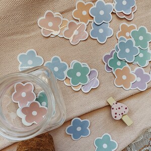 May include: A collection of colorful flower stickers in various shades of blue, green, pink, and orange. The stickers are arranged on a beige fabric background with a glass jar and a wooden heart-shaped clothespin.