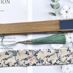 May include: A wooden folding fan with a blue fabric panel and a green tassel. The fan is closed and lying on a white surface with a piece of fabric with a floral pattern.