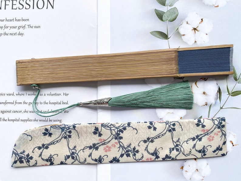 May include: A wooden folding fan with a blue fabric panel and a green tassel. The fan is closed and lying on a white surface with a piece of fabric with a floral pattern.