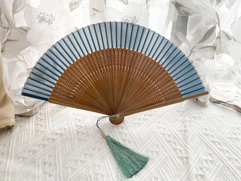 May include: A blue and brown hand fan with a green tassel. The fan is closed and has a wooden frame with a decorative pattern. The fan is on a white surface.